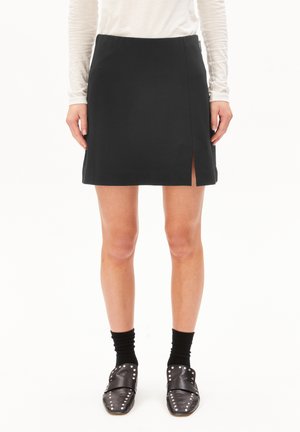 Femme portant une jupe noire avec une fente sur le devant, des chaussures noires à glissière à clous, des chaussettes noires et un haut léger à manches longues, se tenant contre un fond blanc.