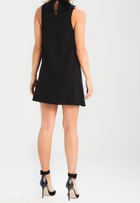 Abito nero senza maniche con dettaglio a frange sul collo, forma a trapezio e lunghezza sopra il ginocchio. Indossato con tacchi neri con cinturino alla caviglia.