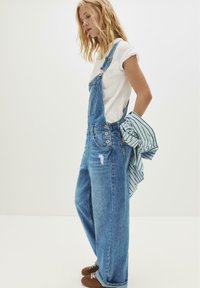 Jonge vrouw in een wit T-shirt en een wijde denim overall houdt een gestreept shirt achter haar rug tegen een effen witte achtergrond.