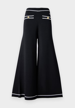 Svarte vide bukser med hvite kantdetaljer, med gullknapper på baklommene og en glatt, teksturert stoffoverflate.