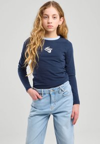 Maglietta a maniche lunghe blu navy con colletto azzurro e logo bianco, abbinata a jeans azzurri a vita alta con tasche laterali.