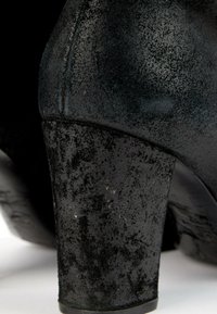 Gros plan sur le talon d'une chaussure en cuir noir montrant une surface usée et texturée avec des éraflures et des plis visibles.
