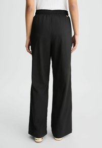 Zwarte wijde broek met een elastische tailleband. Gemaakt van soepel materiaal, met een kleine gouden accent in de buurt van de tailleband.