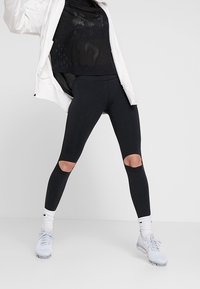 Leggings neri a vita alta con ritagli sulle ginocchia, realizzati in un materiale elasticizzato, abbinati a una maglietta in rete nera e calzature sportive bianche.