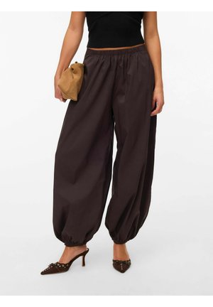 Kvinde iført løse mørkebrune ballonbukser med elastiske manchetter, sort crop top, brunspidse hæle og holder en tan clutch taske.