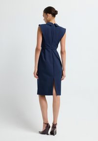 Femme dans une robe sans manches bleu marine, arrivant au genou, avec des épaules larges et une fente dans le dos, portant des chaussures à talons hauts sombres, se tenant devant un fond uni.
