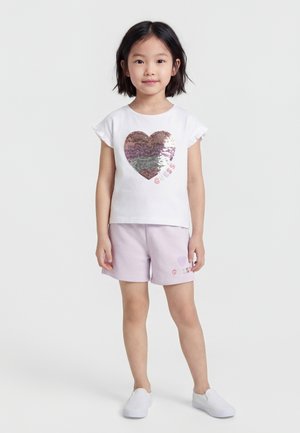 Νέα κοπέλα σε όρθια στάση, φοράει λευκό κοντομάνικο με μεγάλο καρδιά διακοσμημένη με παγιέτες και ανοιχτό μωβ σορτς, λευκά παπούτσια slip-on, ουδέτερο φόντο.