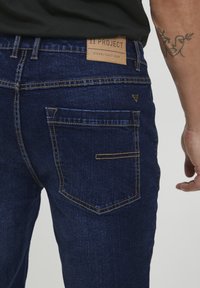 Tamno plave traperice imaju poseban stražnji džep s horizontalnim šavom, žutim šavovima kao akcentima, i kožnom oznakom brenda na struku.