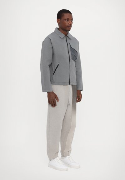 Veste grise avec fermeture éclair à l'avant et poche sur la poitrine, associée à un pantalon de survêtement gris clair. Modèle portant des baskets blanches. Design simple et utilitaire.