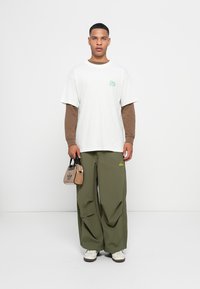 Witte t-shirt met korte mouwen en groen logo, lange bruine mouwen, losse olijfgroene broek en een kleine bruine schoudertas met zwarte accenten.