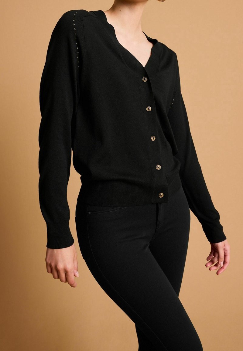 Personne portant un cardigan noir boutonné à manches longues et un pantalon noir ajusté sur un fond beige uni.