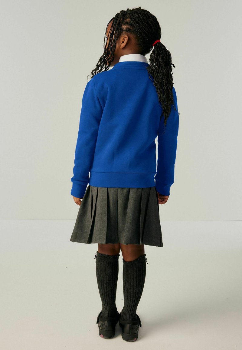 Plava dukserica s dugim rukavima, siva nabirana suknja, crne čarape do koljena s rebrastim uzorkom i crne cipele. Pogled straga.