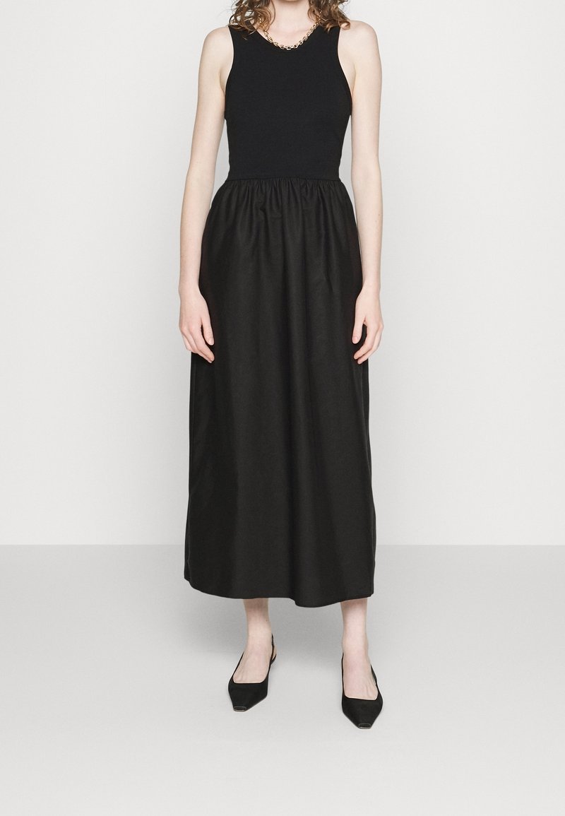 Femme portant un haut noir sans manches et une longue jupe noire avec des chaussures noires pointues, debout devant un fond clair uni.