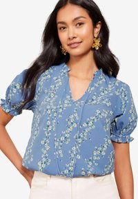 Blusa azzurro chiaro con motivo floreale caratterizzato da margherite bianche. Maniche corte a sbuffo con orli arricciati e scollatura a V. Tessuto morbido.