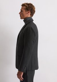 Zwarte textuur blazer met hoge kraag, enkele knoopsluiting en schuine zakken, weergegeven op een model in profiel tegen een effen achtergrond.