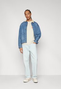 Denim jakk, millel on beež krae, nupulukk ja rinnakott, koos heleda beeži särgi ning heledate siniste lõdvalt istuvate teksadega.