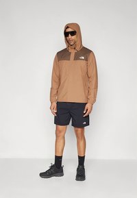 Καφέ και ταμπά αδιάβροχο μπουφάν με κουκούλα, με φερμουάρ στο μπροστινό μέρος και ελαστικά μανίκια, συνδυασμένο με μαύρο σορτς και μαύρα παπούτσια.