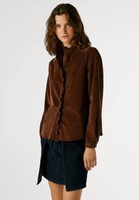 Camisa de botonadura de pana marrón con detalle fruncido en la parte delantera, mangas largas y cuello, combinada con una falda de mezclilla oscura con una abertura lateral.
