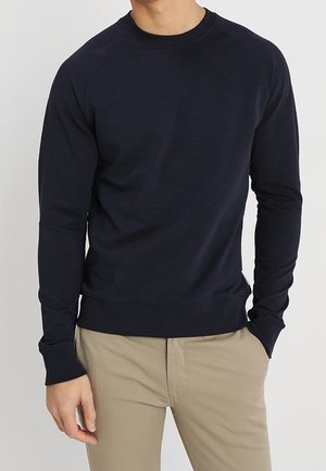 Homme portant un t-shirt cintré bleu marine à manches longues avec un pantalon beige, debout avec une main dans la poche.
