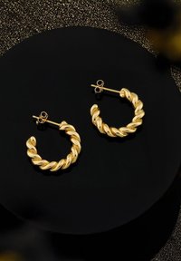 Guldörhängen med en vriden design och glänsande yta, med en stiftbackstängning. Visas på en svart cirkulär bakgrund.
