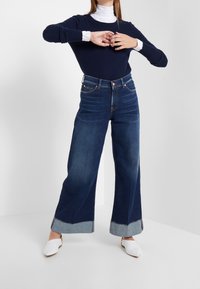 Femme portant un jean bleu foncé à jambes larges et retroussé, un pull marine par-dessus une chemise blanche, et des chaussures blanches à bout pointu, se tenant avec les mains croisées.