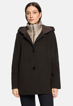 Vrouw met een donkerbruine mantel met capuchon over een lichtbeige doorgestikte jas met een voorrits, staand tegen een effen achtergrond.