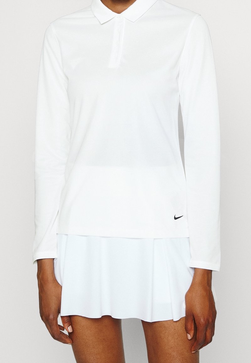 Polo shirt bianco a maniche lunghe con colletto, indossato con una gonna plissettata bianca, caratterizzata da un piccolo logo Nike nero vicino all'orlo.