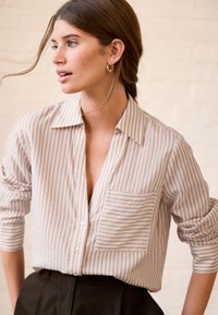 Chemise beige clair à boutons, avec fines rayures verticales blanches, dotée d'une poche poitrine gauche et de manches retroussées, associée à un pantalon foncé.