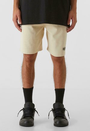 Lys beige bomuldsbukser med sidelommer, med et sort logo på venstre lår, parret med sorte sneakers og ribbede sorte sokker.