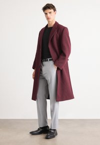 Palton din lână bordeux cu guler cu revere, purtat împreună cu o cămașă neagră ajustată și pantaloni gri deschis. Pantofii negri completează ținuta.
