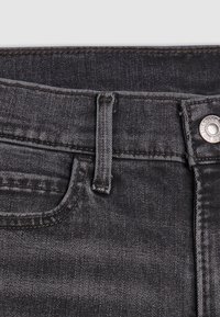 Jeans in denim nero con una superficie strutturata, passanti per cintura e chiusura con bottone in metallo. Cuciture visibili lungo le suture.