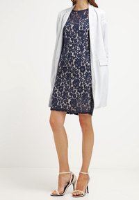 Námořnické krajkové šaty se přiléhavým střihem, vrstveným designem a kulatým výstřihem, doplněné o světle šedou otevřenou bundu a sandály na tenkých podpatcích.