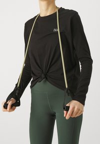 Svart långärmad topp med knuten frontdesign, i kombination med gröna leggings och träningsmotståndsband med svarta handtag och beige remmar.