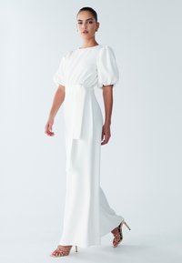 Witte jurk met pofmouwen en een gecentreerde taille, gemaakt van gladde stof. Lang, vloeiend ontwerp dat wordt aangevuld met bandjes van gouden hakken.