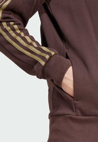 Persona con un sudadera marrón con rayas doradas en las mangas, con la mano colocada de manera casual dentro del bolsillo delantero.