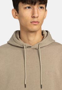 Beige hoodie i mjukt tyg, med kenguruficka, dragsko med silverfärgade ändar och en subtil logo på bröstet.