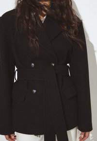 Mustast tekstuurkangast kahe rõngaga mantel, millel on vööd kinnitav vöö, suured taskud ja ees kaks silmapaistvat nuppu.