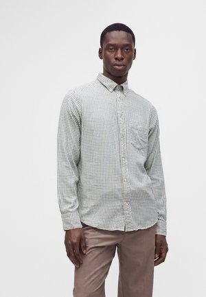 Uomo che indossa una camicia a quadri chiara con bottoni e pantaloni marrone chiaro, in piedi davanti a uno sfondo bianco uniforme.