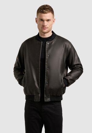 Férfi fekete bőr bomber dzsekiben, fekete ing fölött, kezét zsebébe dugva, szemben áll egy egyszínű világosszürke háttér előtt.