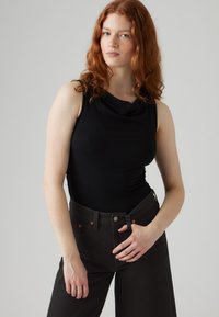 Juodas rankovių palaidinė su drapuotu iškirptu kaklu, derinama su aukšto liemens tamsiai pilkomis džinsais, puoštomis oranžinėmis siūlemis ir užsegama sagomis.