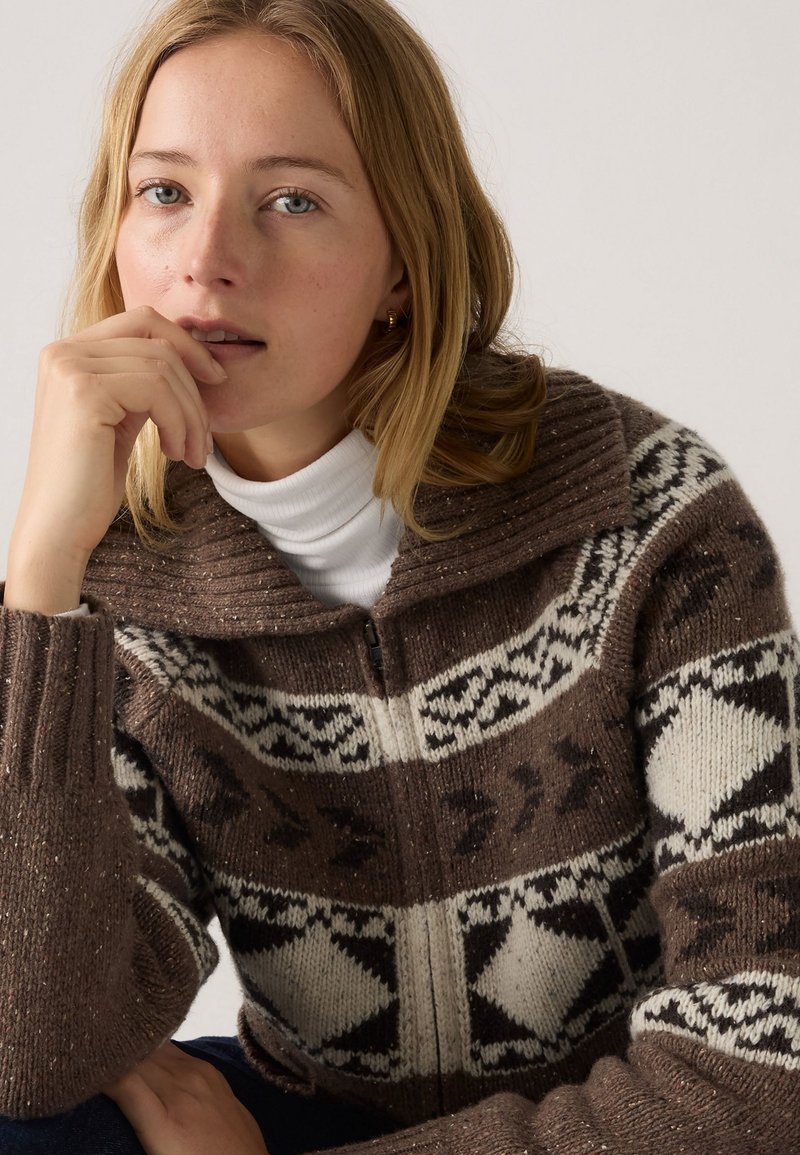 Brun strikket sweater med lynlås foran, der har et fair isle mønster i hvidt og sort. Kraven er ribbet og har en blød tekstur.