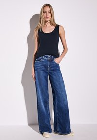 Weite Jeans aus Denim in mittlerem Blauton, mit einem klassischen Fünf-Taschen-Design und sichtbaren Nähten, kombiniert mit einem enganliegenden schwarzen Tanktop.