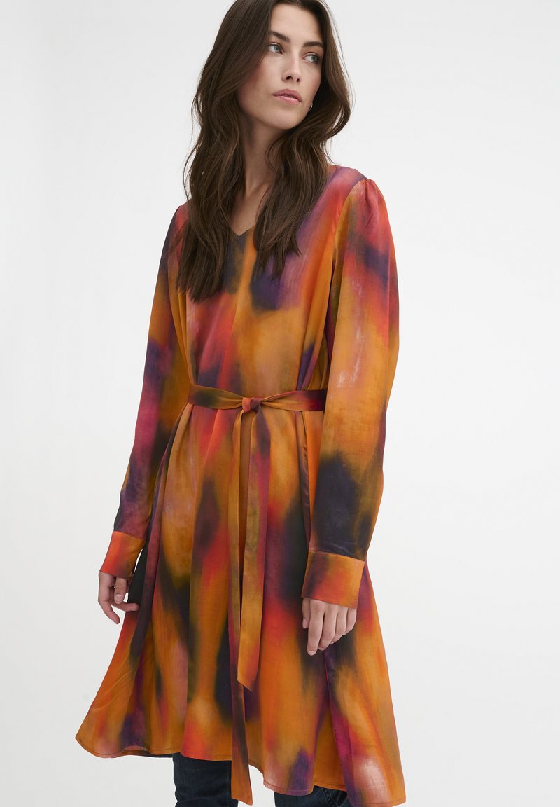 Długa, luźna sukienka z lekkiego materiału, zdobiona intensywnym wzorem tie-dye w kolorze pomarańczowym, czerwonym i purpurowym, z zawiązanym paskiem w talii.