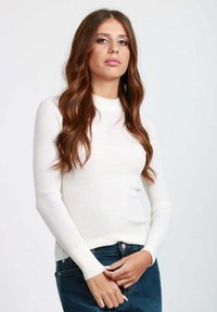 Top lungo a maniche ribattute bianche con collo alto e silhouette aderente. Indossato con jeans blu scuro, che mettono in risalto uno stile casual.