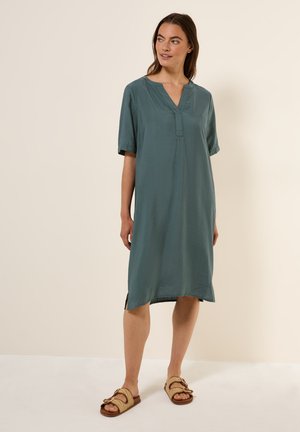 Teal-coloured lyhythihainen mekko, jossa on V-pääntie, hieman löysä malli, sivuhalkiot ja teksturoitu kangas. Yhdistetty ruskeisiin sandaaleihin, joissa on solkiyksityiskohta.