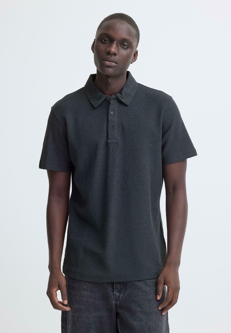Zwart geribd poloshirt met kraag en een drieknopenbies, gecombineerd met donkere denim shorts, weergegeven tegen een neutrale achtergrond.