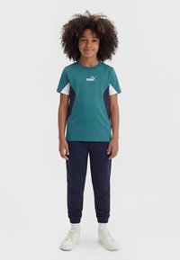 Παιδί με σγουρά μαλλιά που φοράει τιρκουάζ μπλουζάκι Puma, ναυφί μπλέ φόρμα, λευκές κάλτσες και λευκά αθλητικά παπούτσια, στέκεται μπροστά σε απλό ανοιχτό φόντο.