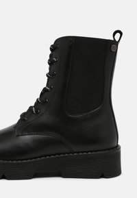 Botte de combat en cuir noir à lacets avec semelle épaisse à rainures, panneau latéral élastique et languette à l'arrière.