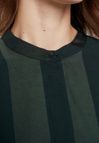 Tummanvihreä paita, jossa on pystysuorat raidat, pyöreä kaulus ja yksi musta nappi yksityiskohtana kaula-aukossa. Materiaalina sileä kangas.