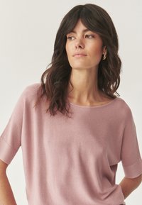 Lichtroze gebreide top met een ruime halslijn en korte mouwen, met een zachte textuur en een relaxte fit. Zilveren hardware-accenten zichtbaar.
